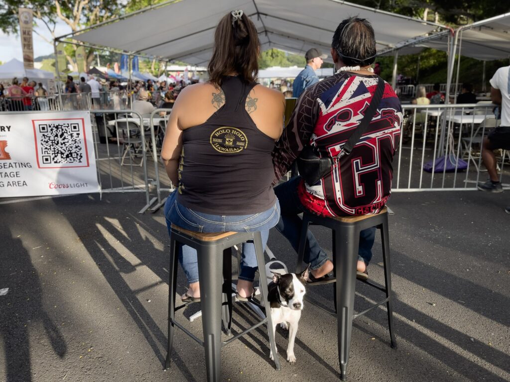 カネオヘ・ファーマーズマーケットのライブステージ前で、「Hawaiʻi ’78」を聴くハワイアンのカップルと、その足元に座る犬。